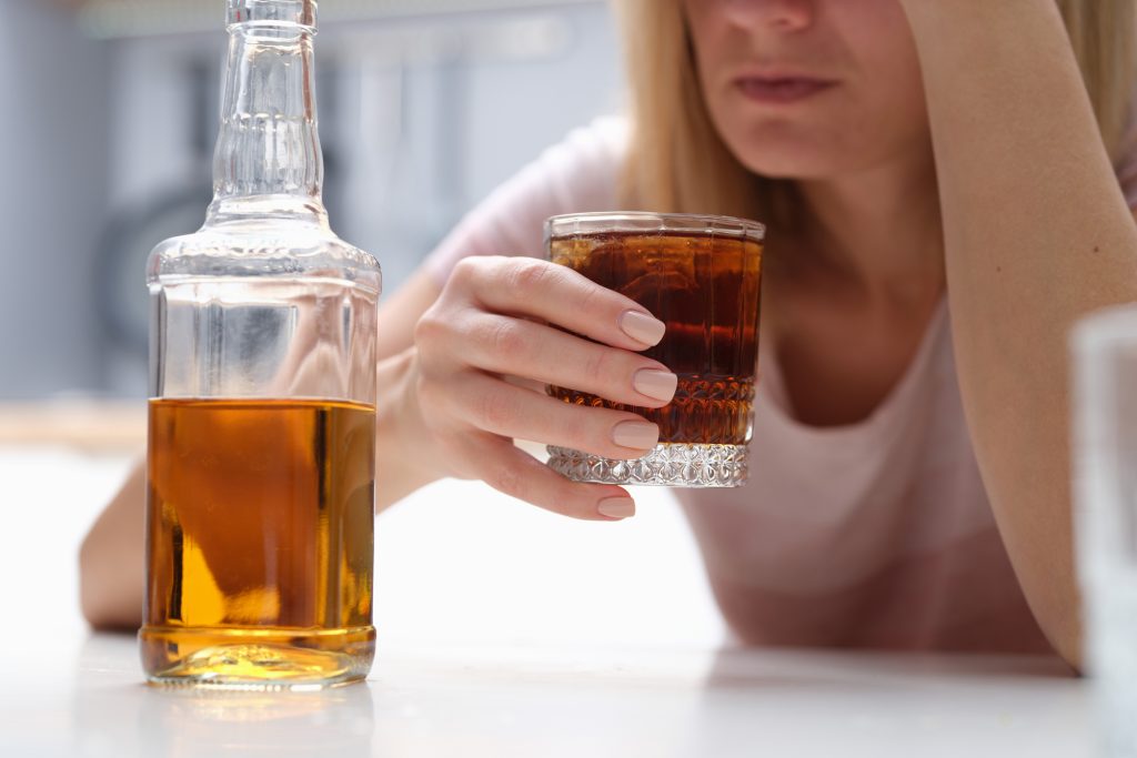 a woman holding a whiskey glass filled to the brim. The glass whiskey bottle is sitting next to her. She looks depressed and has her arm and hand on her head.