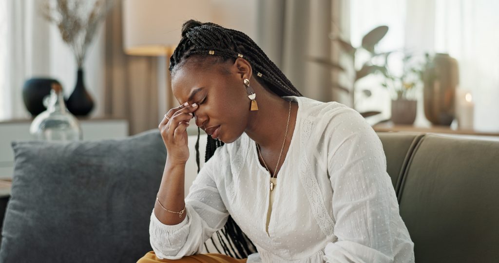 A young black woman sitting on her couch looking stressed out. Her hand is holding her face.